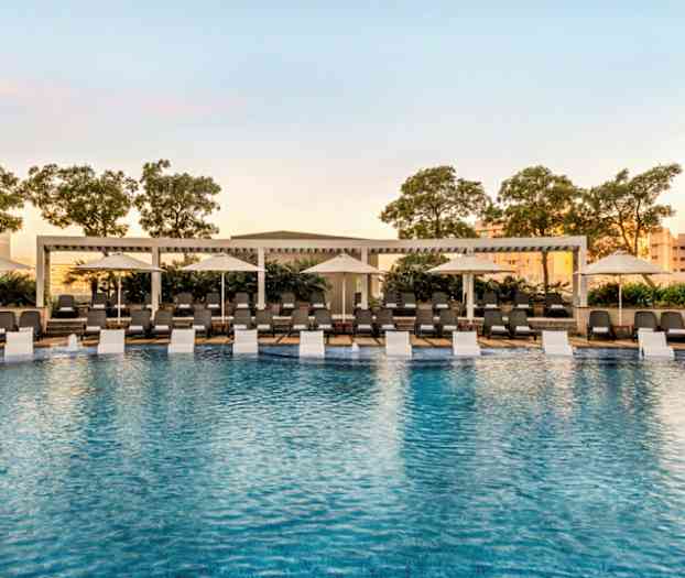 Utomhuspool omgiven av solstolar och parasoller med träd i bakgrunden. (AI-genererad text)