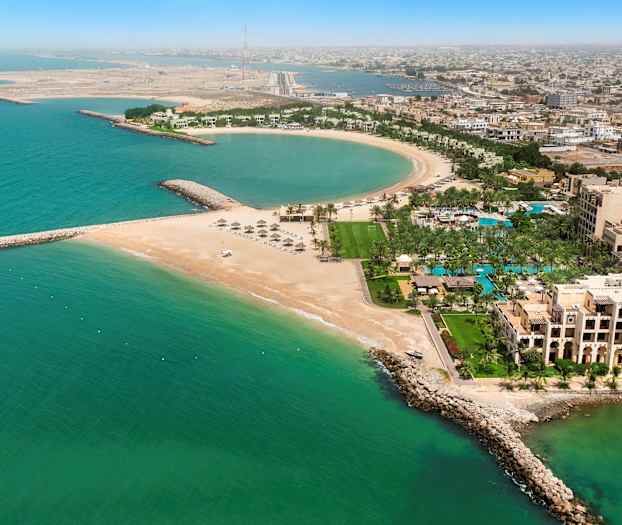 Ett flygfoto av en kuststad med en halvrund strand, byggnader, palmer och turkost havsvatten. (AI-genererad text)