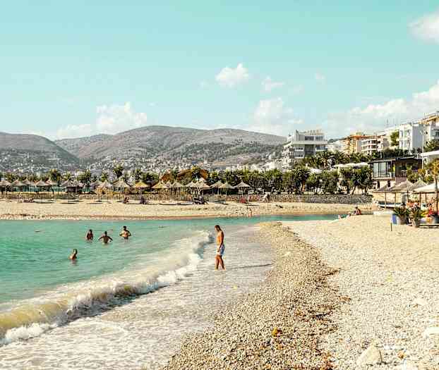 Strand med parasoller och badande människor i klart vatten. Byggnader och berg i bakgrunden. (AI-genererad text)