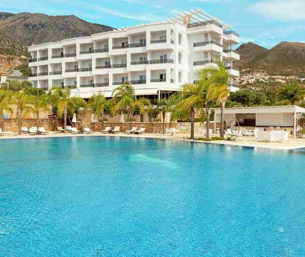 En utomhuspool framför ett hotell byggt i en modern vit design, omgiven av palmer och berg i bakgrunden. (AI-genererad text)