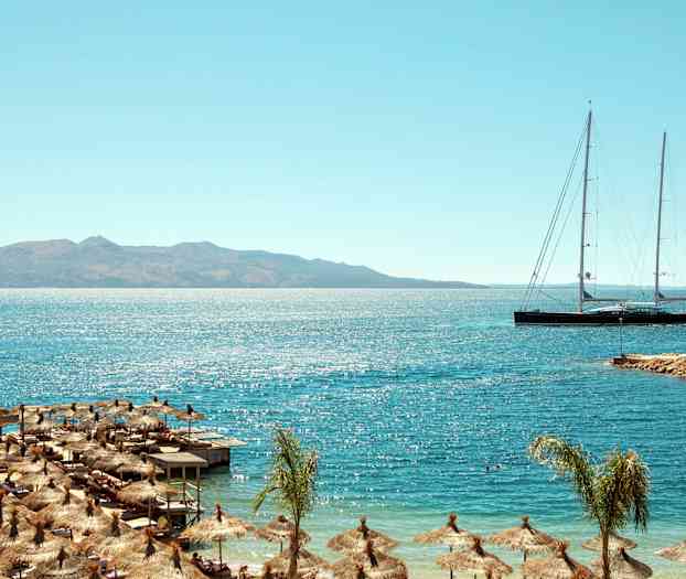 Strand med parasoller och lång segelbåt på havet med berg i bakgrunden. (AI-genererad text)