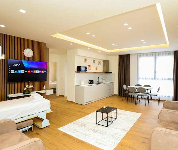 Ett modernt hotellrum med öppen planlösning inkluderande en bäddsoffa, TV, köksområde, matbord och stora fönster med gardiner. (AI-genererad text)