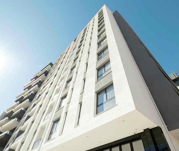 Bilden visar en modern byggnad med flera våningar, fasaden är ljus och har många fönster. En klar blå himmel och direkt solljus syns. (AI-genererad text)