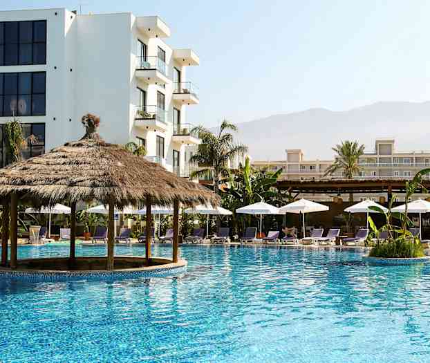 En pool med en halmtakspaviljong omgiven av solstolar och parasoller, med flerbyggnadskomplex i bakgrunden. (AI-genererad text)