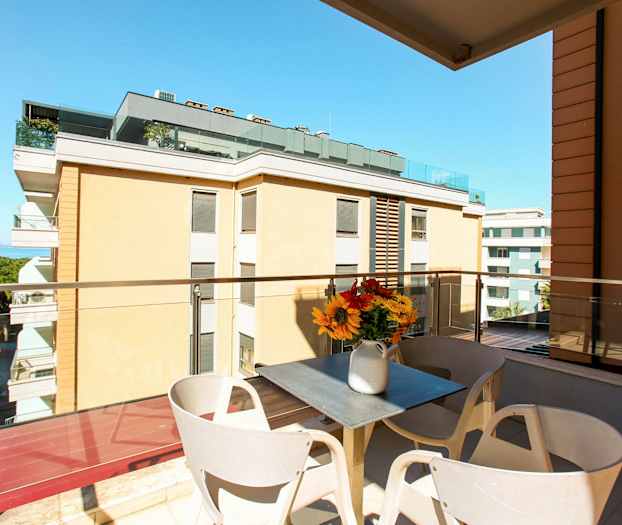 En balkong med utsikt över byggnader och havet. På balkongen finns ett bord med en vas med blommor. (AI-genererad text)