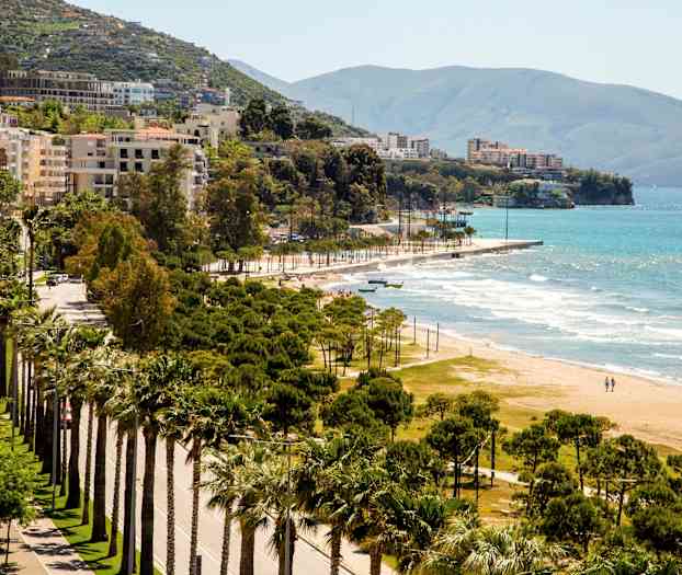 Vy över en kustlinje med sandstrand, grönskande träd och byggnader i bakgrunden. (AI-genererad text)