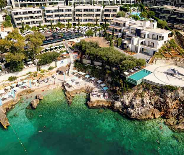 Flygbild av ett kusthotell med pool, helikopterplatta och strand mot turkost vatten. (AI-genererad text)