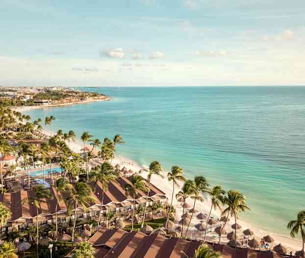 Flygfoto av en strand med palmer, hotellbyggnader och parasoller nära klart blått hav. (AI-genererad text)