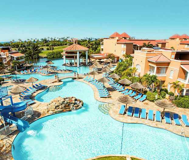 Bild av ett semesterhotell med poolområde, solstolar och parasoller. Byggnader omgärdar området och gröna palmer syns i bakgrunden. (AI-genererad text)