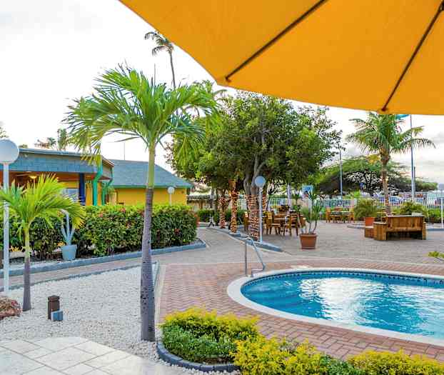 Utomhusområde vid en pool omgivet av växter, palmer, och utomhusbord. I förgrunden skymtar ett gult parasoll. (AI-genererad text)