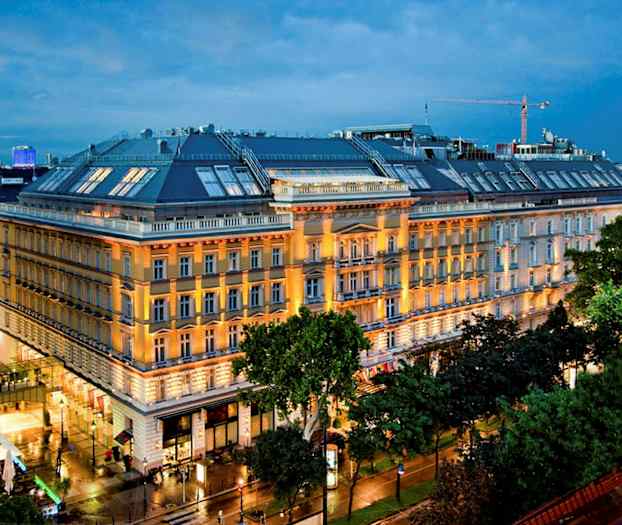 En stor byggnad med flera våningar belyst av fasadbelysning, omgiven av grönområden och städer i bakgrunden. (AI-genererad text)