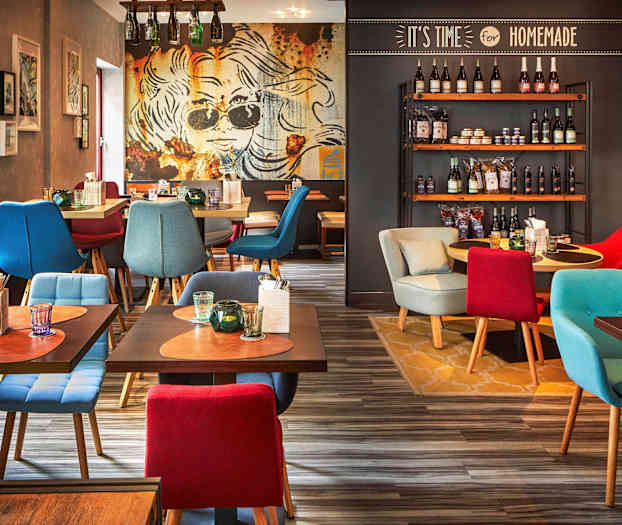 Ett restauranginteriör med färgglatt sittande möbler, dekorativa bord med dryck och bestick samt en vägg med hyllor fyllda med olika produkter. (AI-genererad text)