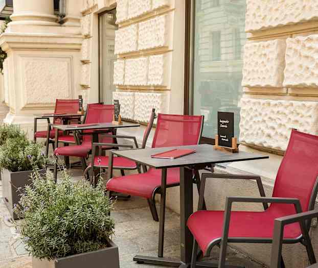 Utomhus bord och stolar med röda sittdynor är placerade längs en gångväg vid en byggnad. (AI-genererad text)