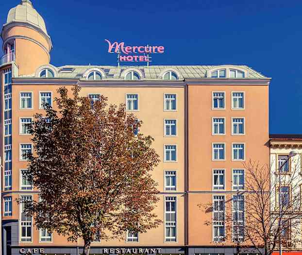 Exteriören av ett hotell med namnet Mercure, med en beige fasad, många fönster och ett högt torn upptill. (AI-genererad text)