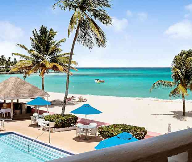 Ett tropiskt strandlandskap med ett turkost hav, vita sandstränder, palmer och en pool i förgrunden. Parasoll och utemöbler är placerade nära poolen. (AI-genererad text)