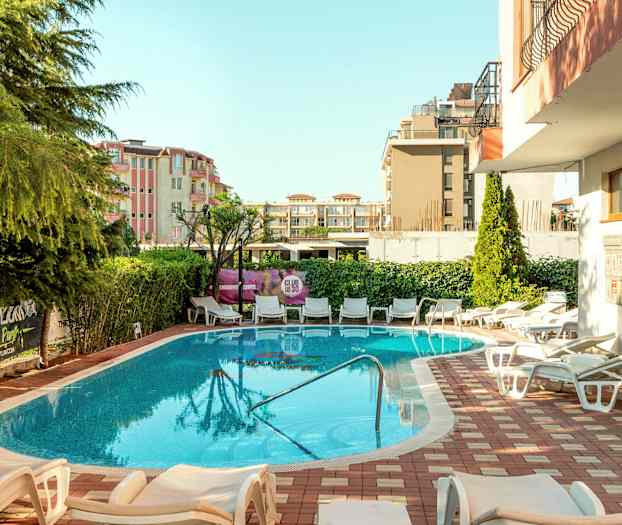 En utomhuspool omgiven av solstolar och grönska med byggnader i bakgrunden. (AI-genererad text)