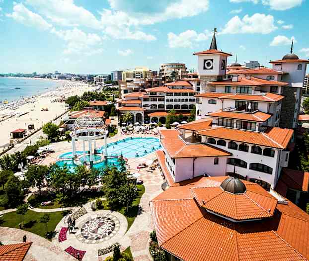 En resort med pool, omgärdad av byggnader med röda tak, ligger vid en strand med parasoller och solstolar. (AI-genererad text)