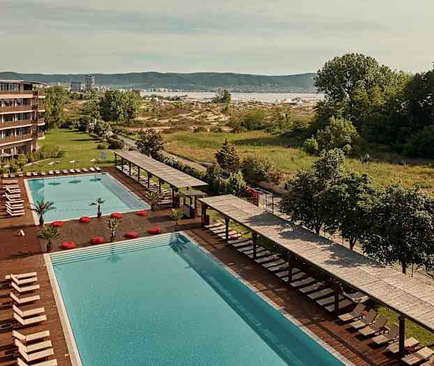 Stor pool med solstolar och parasoller vid sidan av ett hotell, med grönska och landskap i bakgrunden. (AI-genererad text)