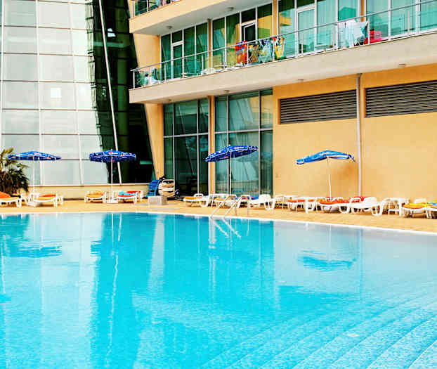 En utomhuspool med solstolar och parasoller vid kanten, belägen framför en byggnad med glasrutor och balkonger. (AI-genererad text)