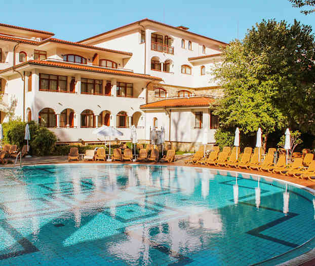 En pool framför en vit byggnad med röda tak, omgiven av solstolar och gröna träd. (AI-genererad text)