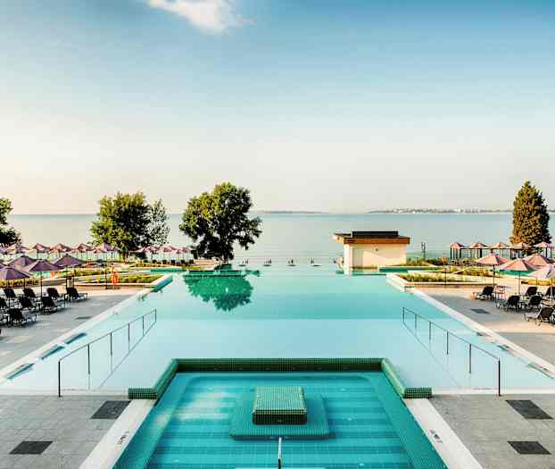 En utsikt över en pool med solstolar och parasoller utspridda runt omkring, samt havet och träd i bakgrunden. (AI-genererad text)