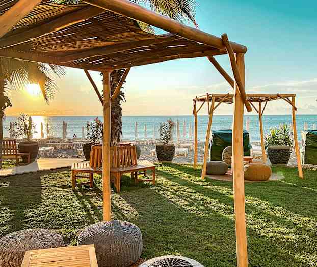 Utemöbler under träskärmar nära stranden vid solnedgång. (AI-genererad text)