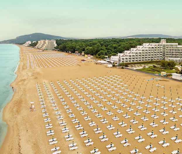 Flygbild av en stor sandstrand med rader av solstolar och parasoller, samt byggnader och grönska i bakgrunden. (AI-genererad text)