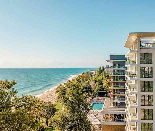 En vy från höjd över havskusten med ett hotell på höger sida och stranden omgiven av träd. (AI-genererad text)