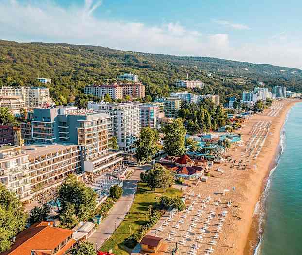 Foto av en kuststad med sandstrand, parasoller och byggnader intill grönskande kullar. (AI-genererad text)