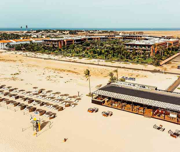 Flygvy av en sandstrand med parasoller och solstolar, samt en strandrestaurang med bord och stolar, omgivet av resortbyggnader och gröna växter. (AI-genererad text)