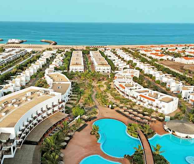 Flygbild av ett hotellområde med pooler, byggnader och strand i bakgrunden. (AI-genererad text)