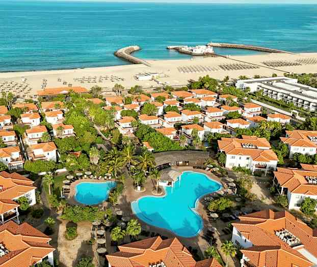 Flygvy över ett semesterområde med vita hus med orangea tak, swimmingpool och en sandstrand vid havet. (AI-genererad text)