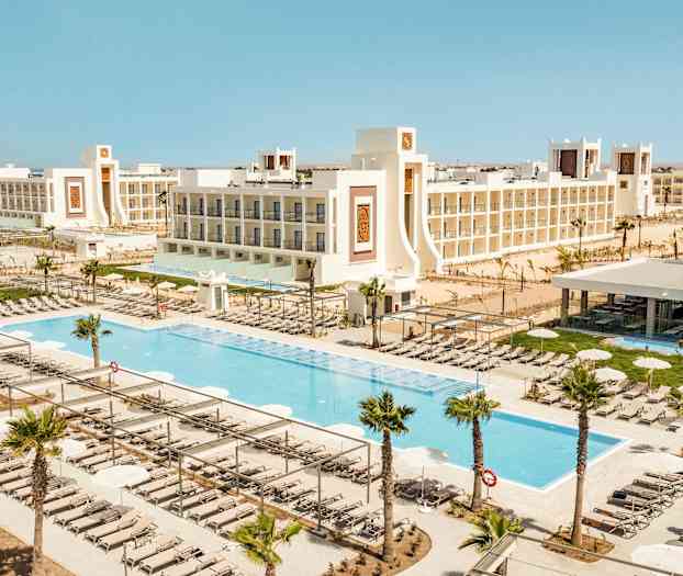 En vy över en semesteranläggning med pooler, solstolar och omgivande vita byggnader. (AI-genererad text)