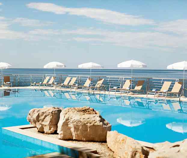 En utomhuspool med solstolar och parasoller i bakgrunden, havet syns bortom poolområdet. (AI-genererad text)