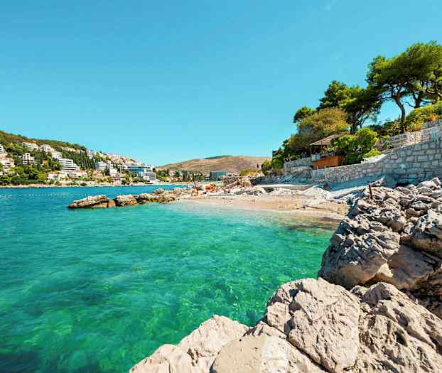 En stenig strand vid ett klart turkost hav med byggnader och träd i bakgrunden under klarblå himmel. (AI-genererad text)