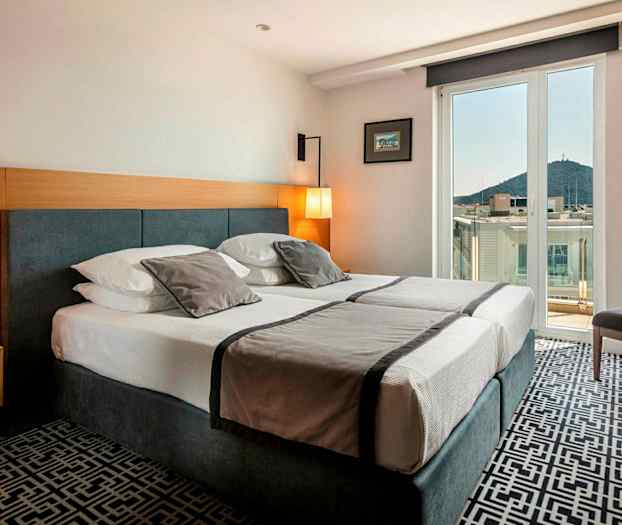 Hotellrum med dubbelsäng, ljus inredning och balkong med utsikt över stad och berg. (AI-genererad text)