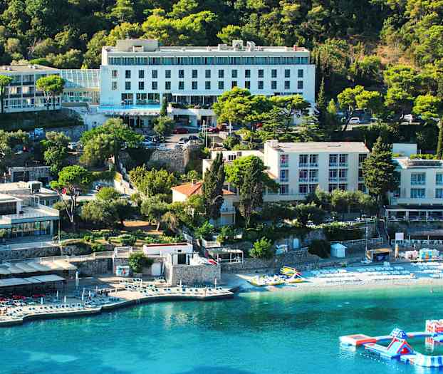 En vy över en hotellbyggnad vid en kust med solstolar på stranden och en flytande lekplats i vattnet. (AI-genererad text)