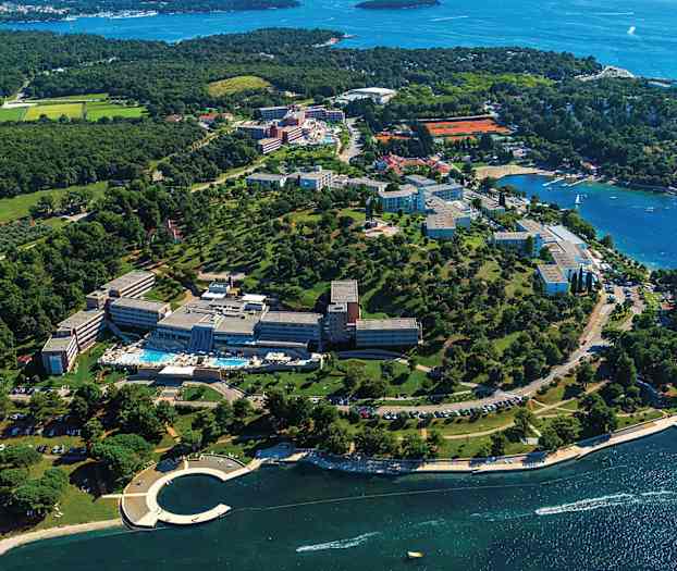 Flygfoto över en semesteranläggning vid kusten med gröna omgivningar och vy över havet. (AI-genererad text)