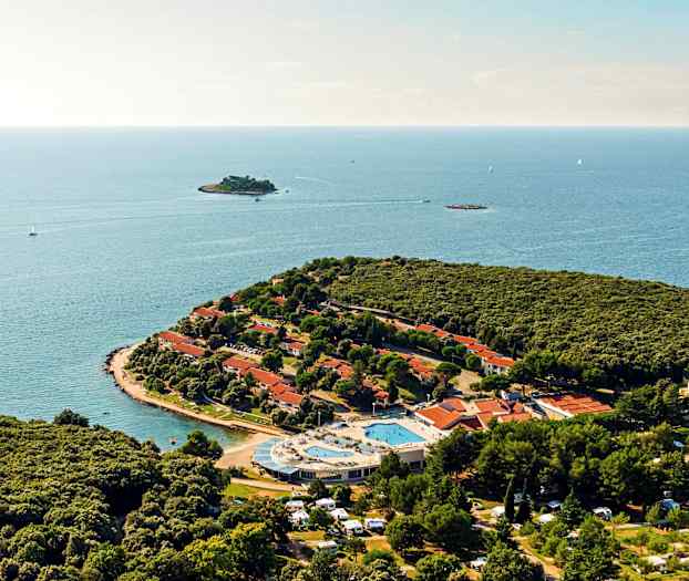 Flygvy över en halvö med tät grönska, röda takhus, en poolområde samt det omkringliggande havet. (AI-genererad text)