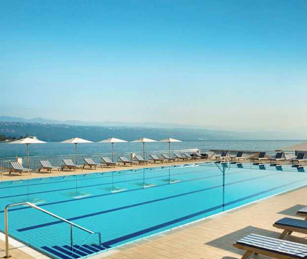 En utomhuspool med solstolar och parasoller vid kanten. I bakgrunden syns havet och berg. (AI-genererad text)