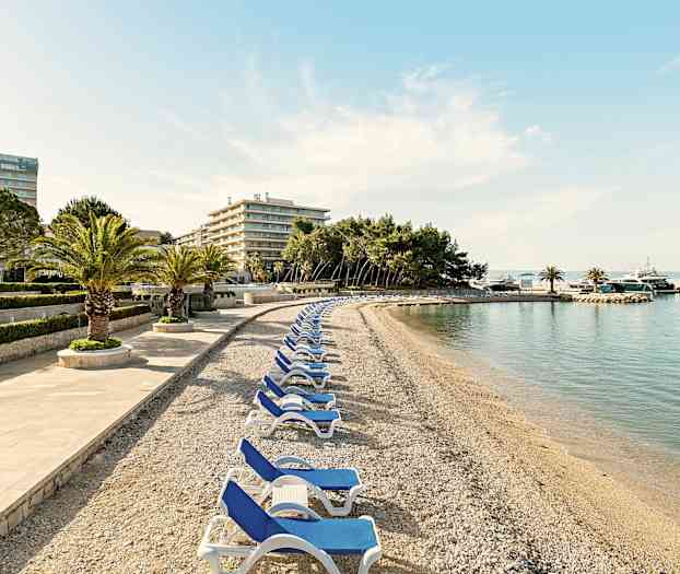 En stenstrand med solstolar och palmer, omgiven av hotellbyggnader och träd. (AI-genererad text)