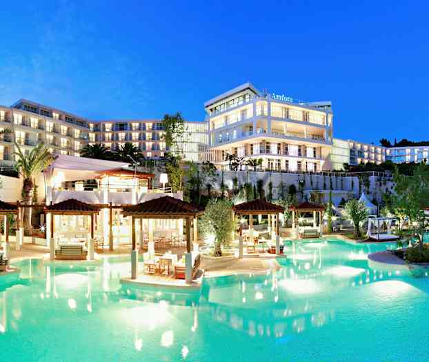 Ett upplyst hotellkomplex med flera våningar vid en pool, omgiven av trädgårdar och palmer, i skymning. (AI-genererad text)