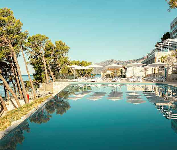 Utsikt över en infinitypool med solstolar och parasoller vid kanten, omgiven av träd och byggnader. (AI-genererad text)