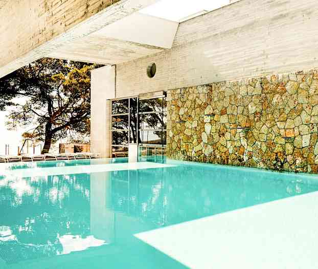 Inomhuspool med stenvägg och utsikt över träd utanför. (AI-genererad text)