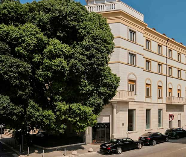 En byggnad med flera våningar och klassisk stil med bågformade fönster och fasad i ljus färg. Framför byggnaden står bilar parkerade bredvid en stor grönskande träd. (AI-genererad text)