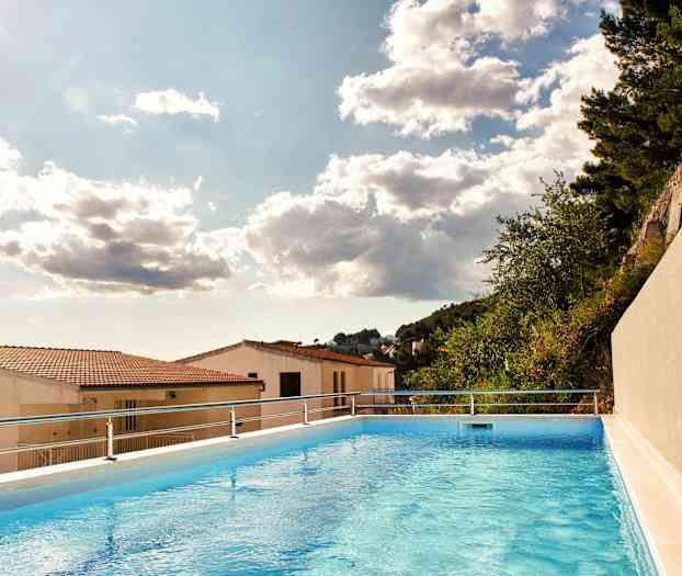 Bild på en utomhuspool med en vy över hustak och träd under en molnig himmel. (AI-genererad text)