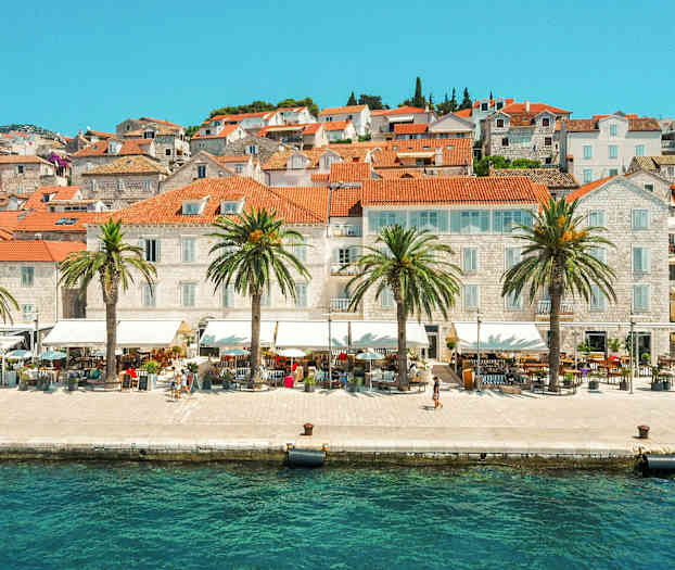 Bild på en stad vid vattnet med tegelröda tak och palmer längs strandpromenaden. (AI-genererad text)