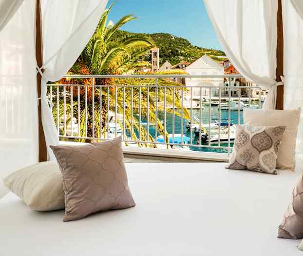 En säng med dekorativa kuddar under en vit baldakin med utsikt över en palm och en hamn. (AI-genererad text)