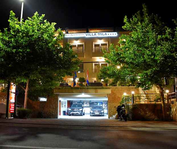 Hotellbyggnad med upplyst entré och parkeringsgarage, omgiven av träd, på natten. (AI-genererad text)