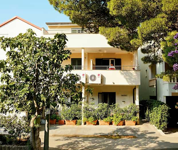 Tvåvåningshus med balkonger och luftkonditioneringsenheter, omgiven av grönska och blommor. (AI-genererad text)